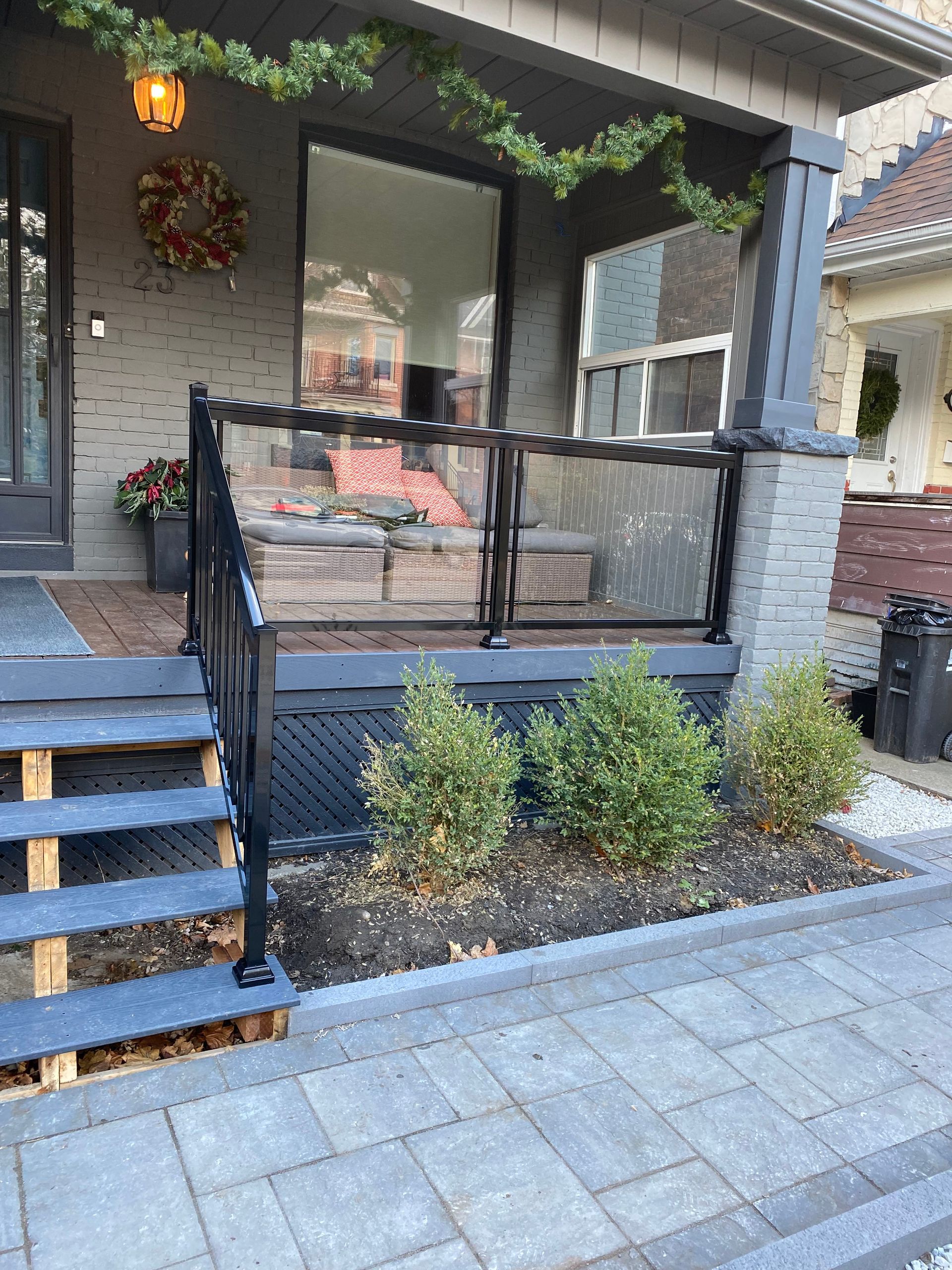 Front porch with glass railing, steps, and greenery. Black railing, grey stone.