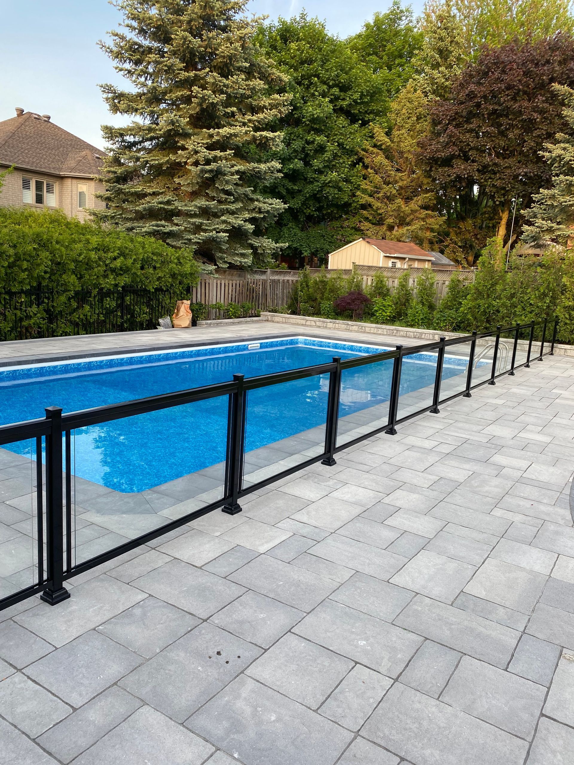A backyard pool with a black fence on a gray stone patio. Green trees and a house are in the background.