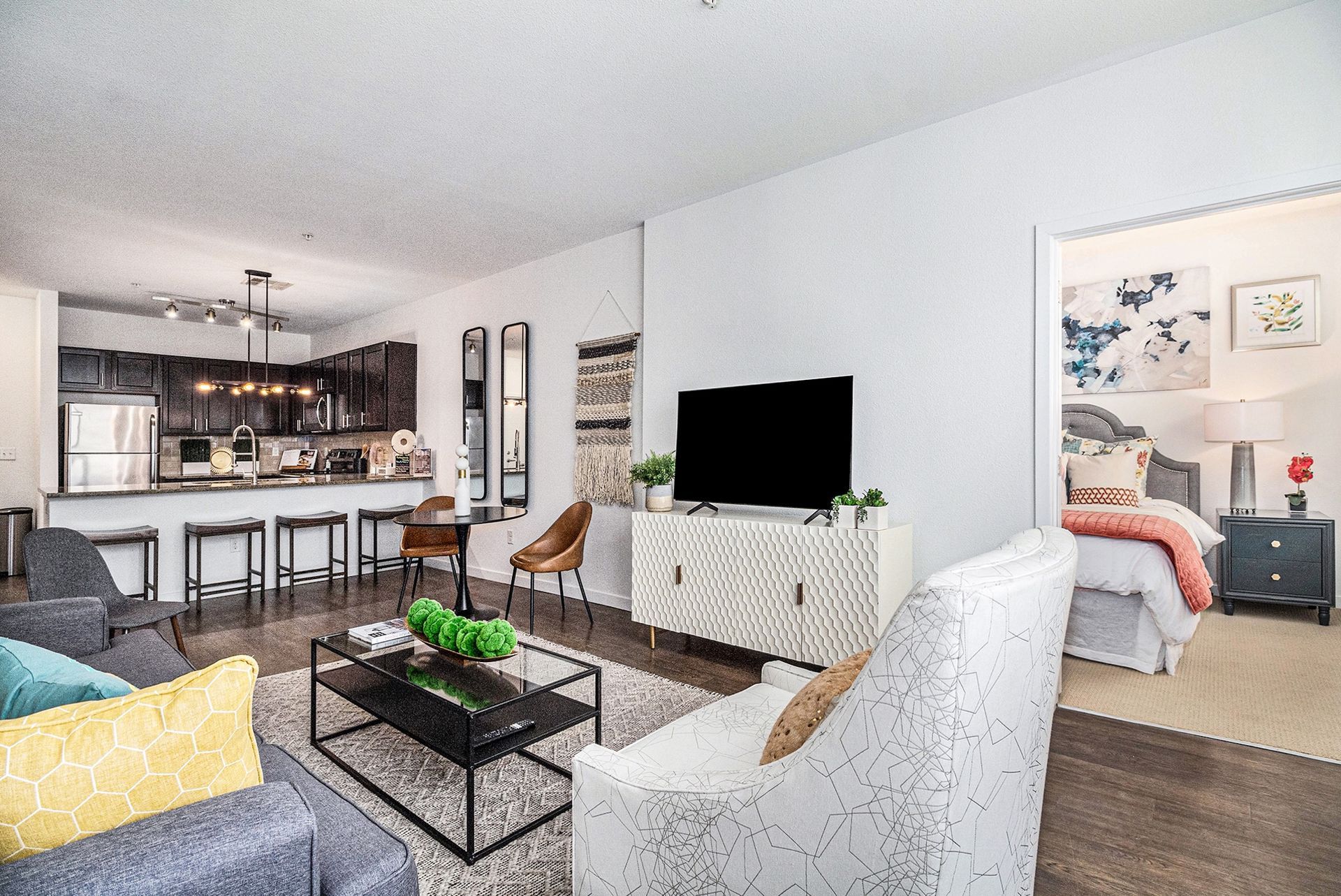 Open-plan living room with kitchen bar, TV, and white armchair leading to a bedroom.