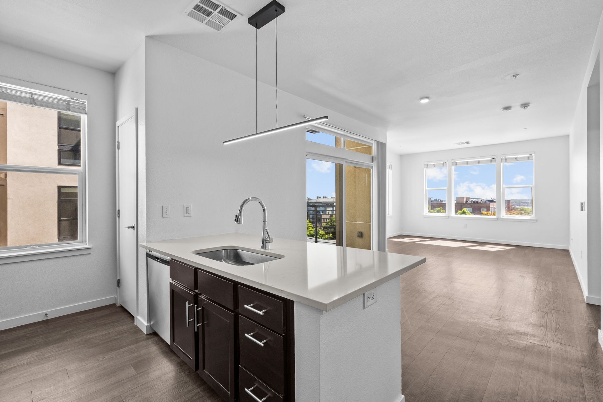 Bright open apartment kitchen with dark cabinets, white counters, and a view into a carpeted living room.