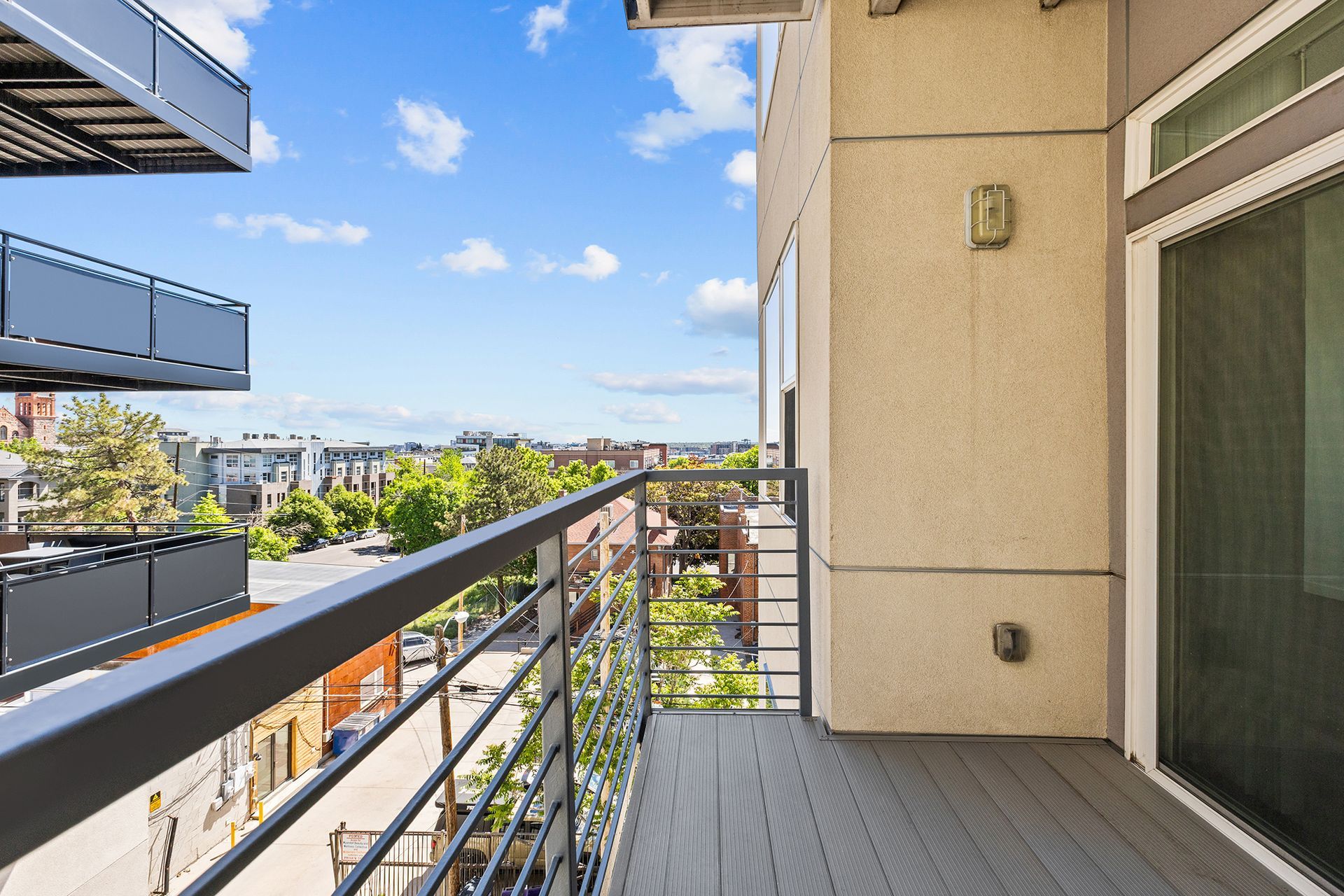 Apartment balcony with gray railing, tan wall, and city view under a blue sky