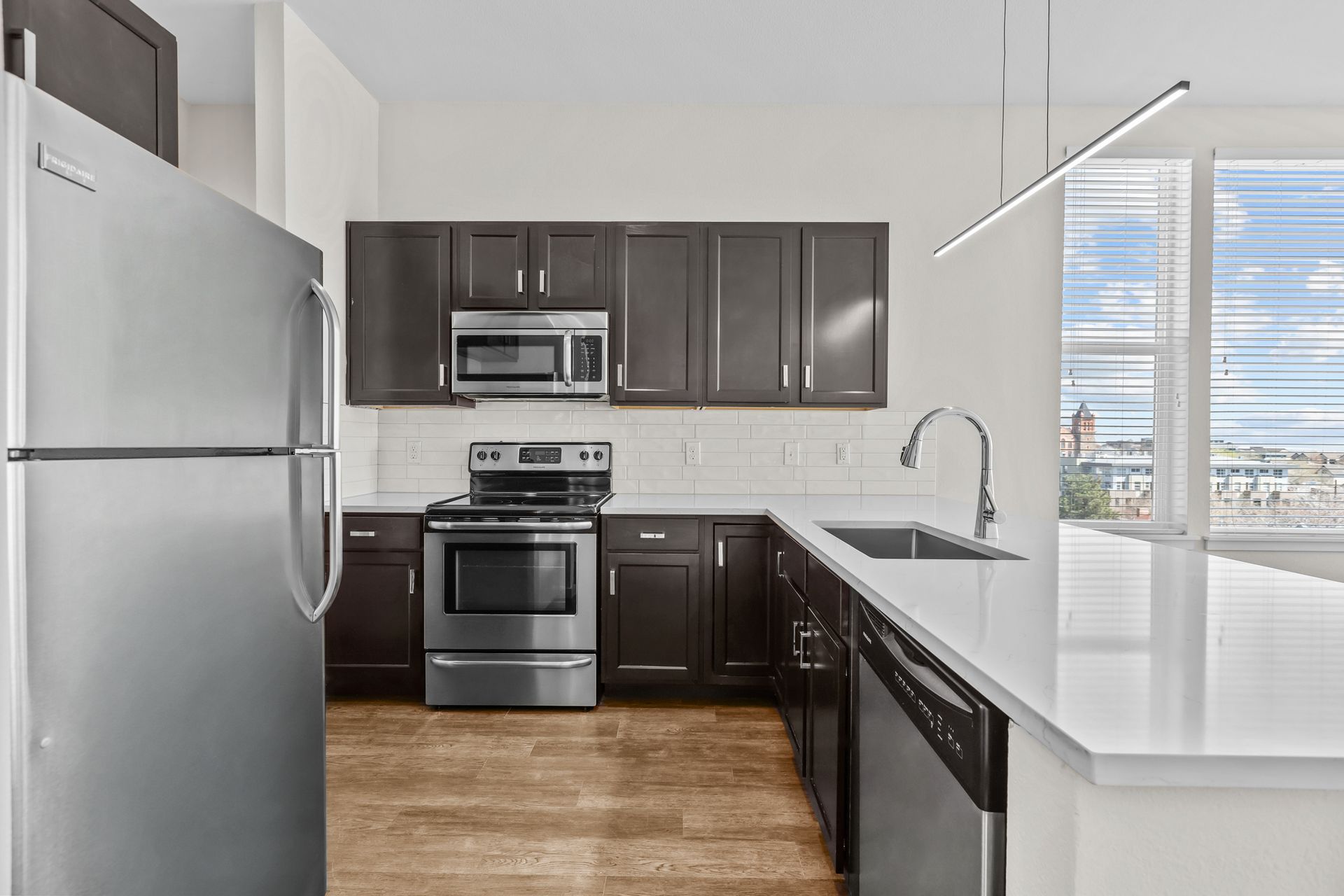 Modern kitchen with stainless steel appliances, dark cabinets, white countertops, and a large window.