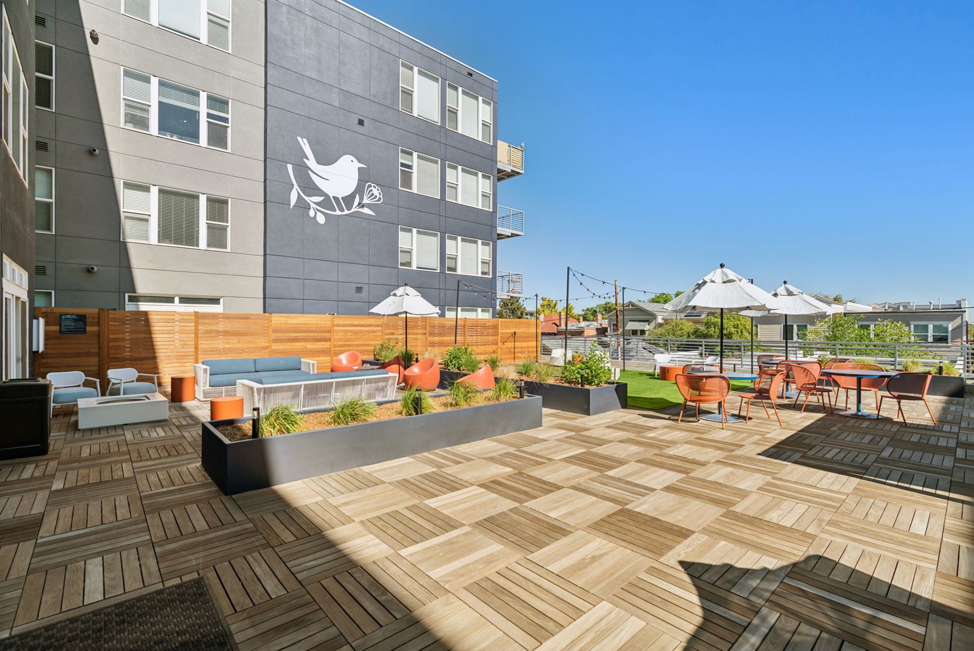 Rooftop patio with orange seating, white umbrellas, potted plants, and a gray building wall
