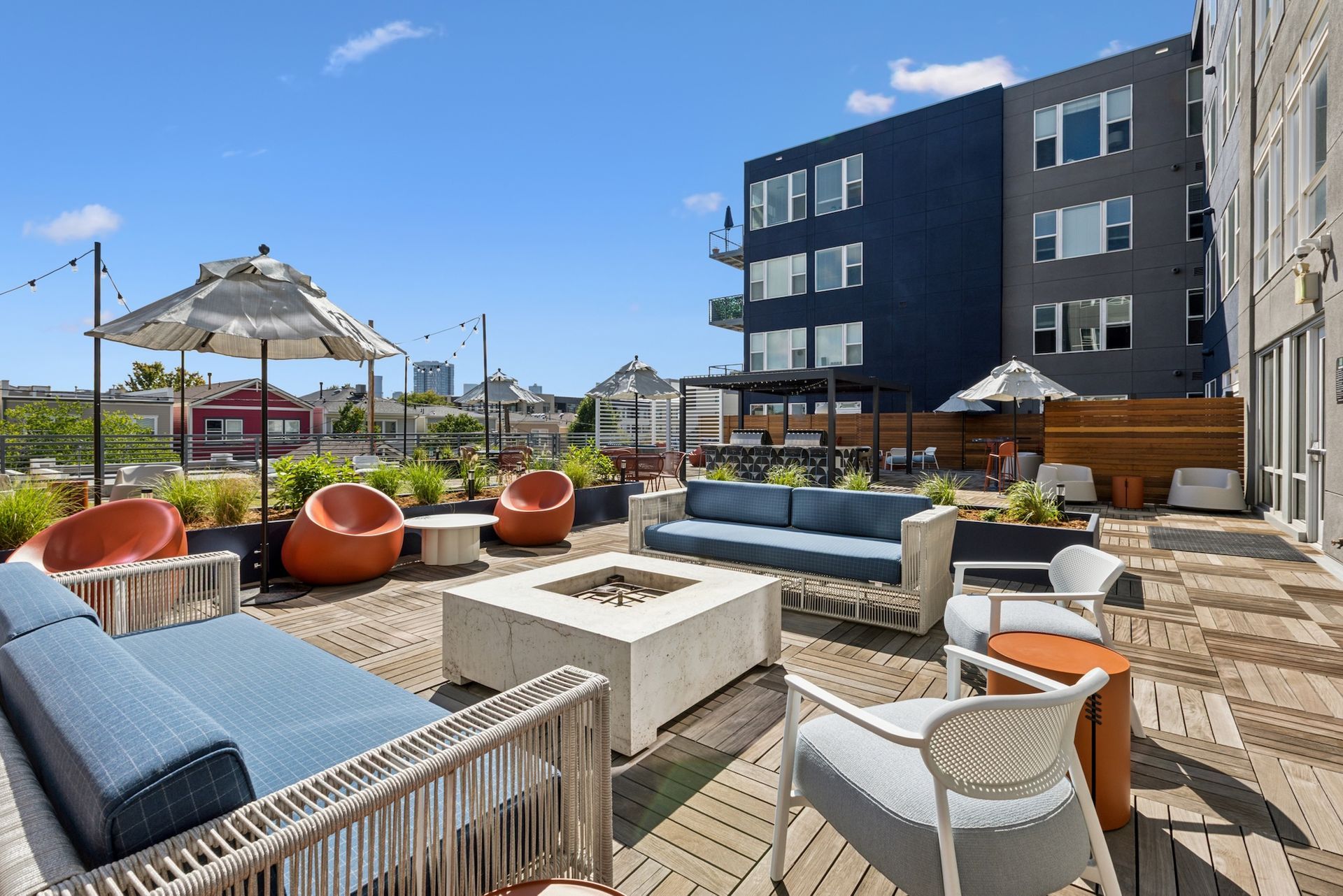 Rooftop patio with blue couches, orange chairs, and fire pit. City buildings in background. Sunny day.