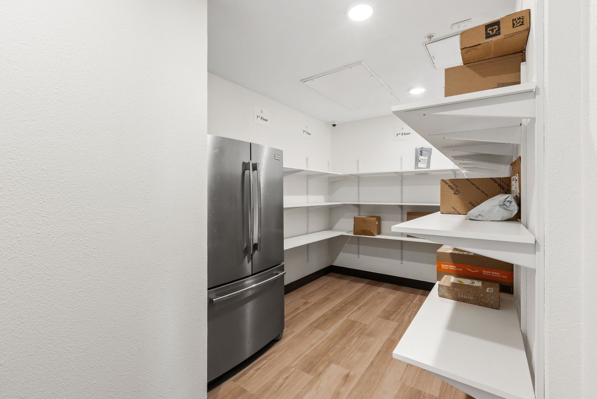 Modern white kitchen with stainless steel fridge, wooden floors, and open shelving, boxes on shelves