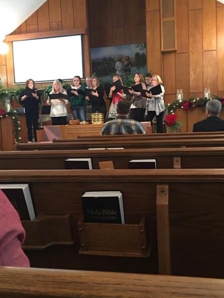 Choir singing on a church stage with a piano, wood-paneled walls, and pews in the foreground