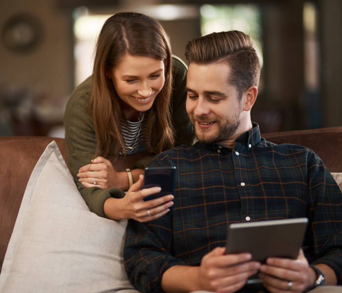 Two people smiling on a couch, looking at a smartphone and tablet in a cozy living room.