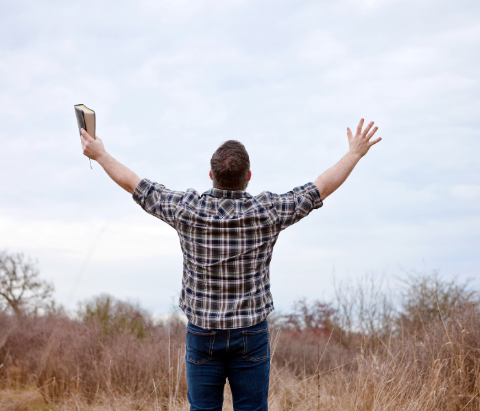 Person standing in a field with arms raised, holding a phone against a cloudy sky.