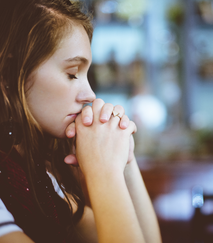 Person in profile with eyes closed, hands clasped in prayer in a softly lit indoor setting