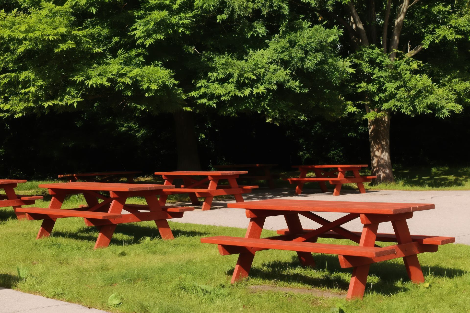 Empty red picnic tables sit on rich green grass under large trees in park.