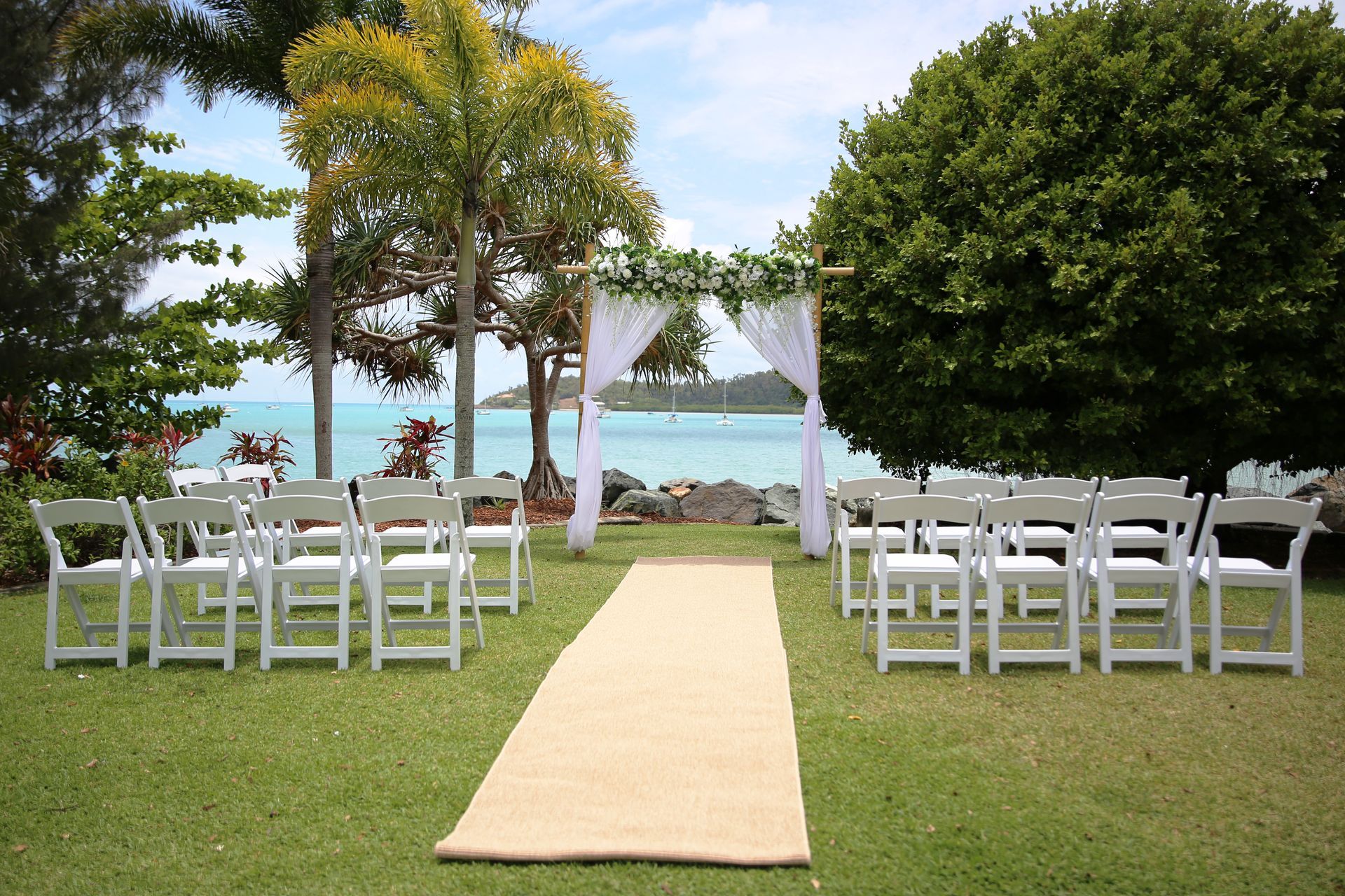 Outdoor wedding with chairs facing an arch draped in white fabric, overlooking water and greenery.