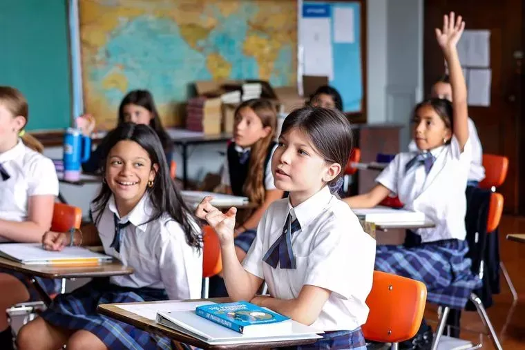 Classroom of students in uniform; girl raises hand; map on wall.