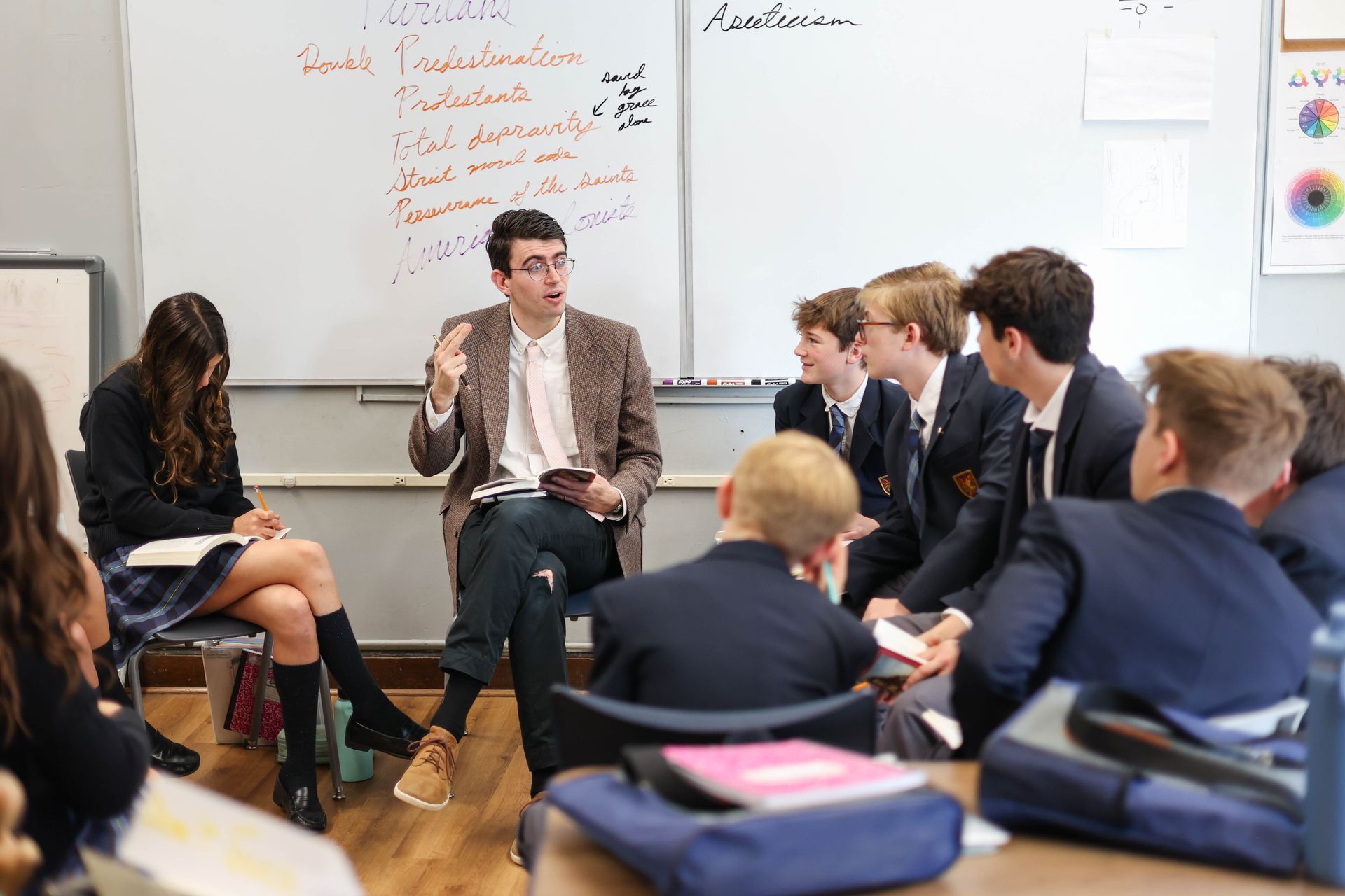 Teacher gesturing while speaking to students in a classroom; whiteboard with notes in background.