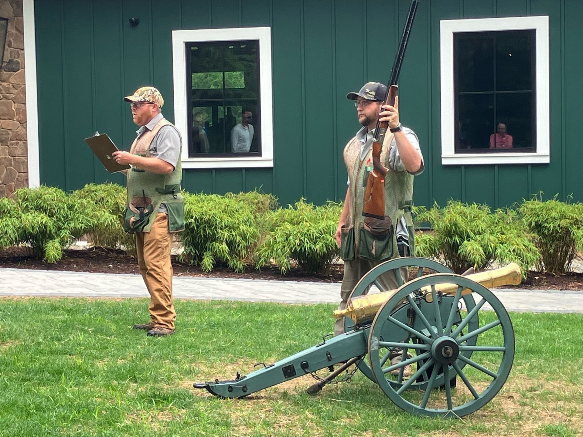 Two men in vests, one holding a shotgun, the other a clipboard, stand near a cannon on a lawn.