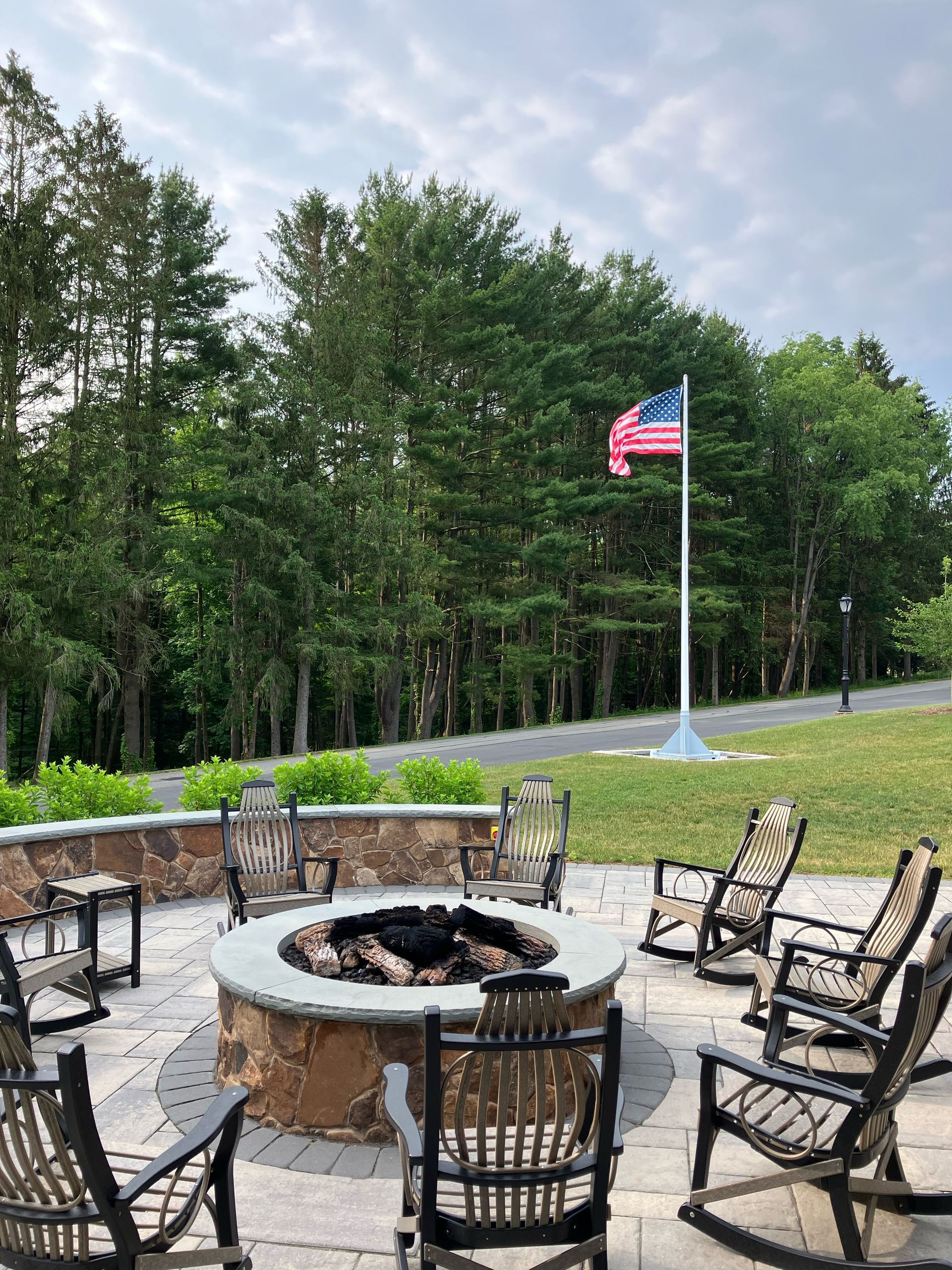 A stone fire pit surrounded by rocking chairs, with a US flag waving in the background.