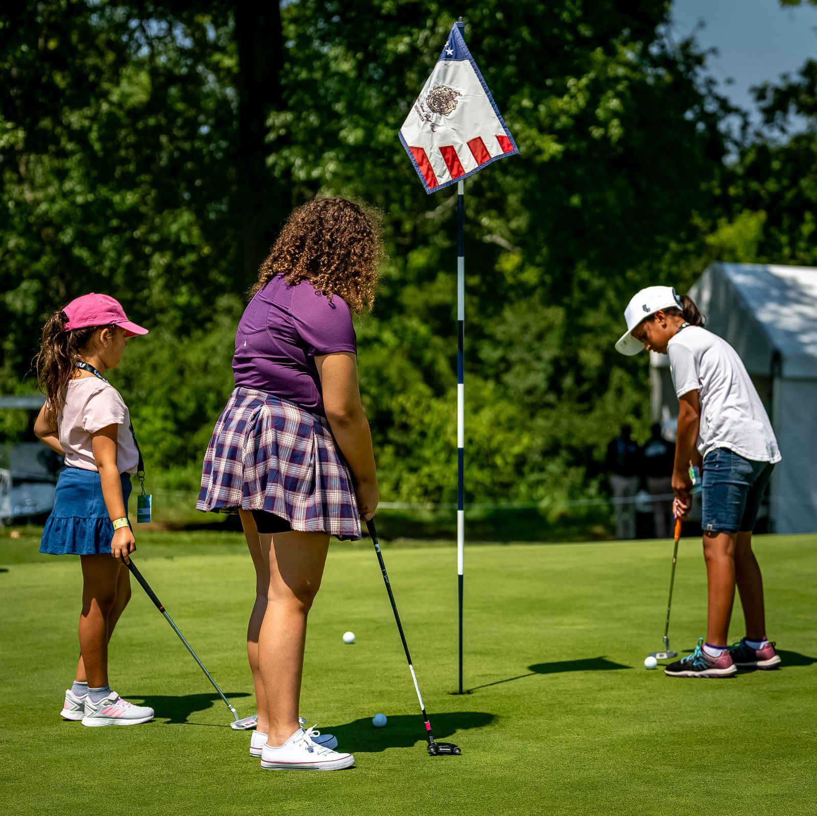 Three people putting golf balls on a green. A woman in purple and two girls wearing hats are focused on their swings. American flag in background.