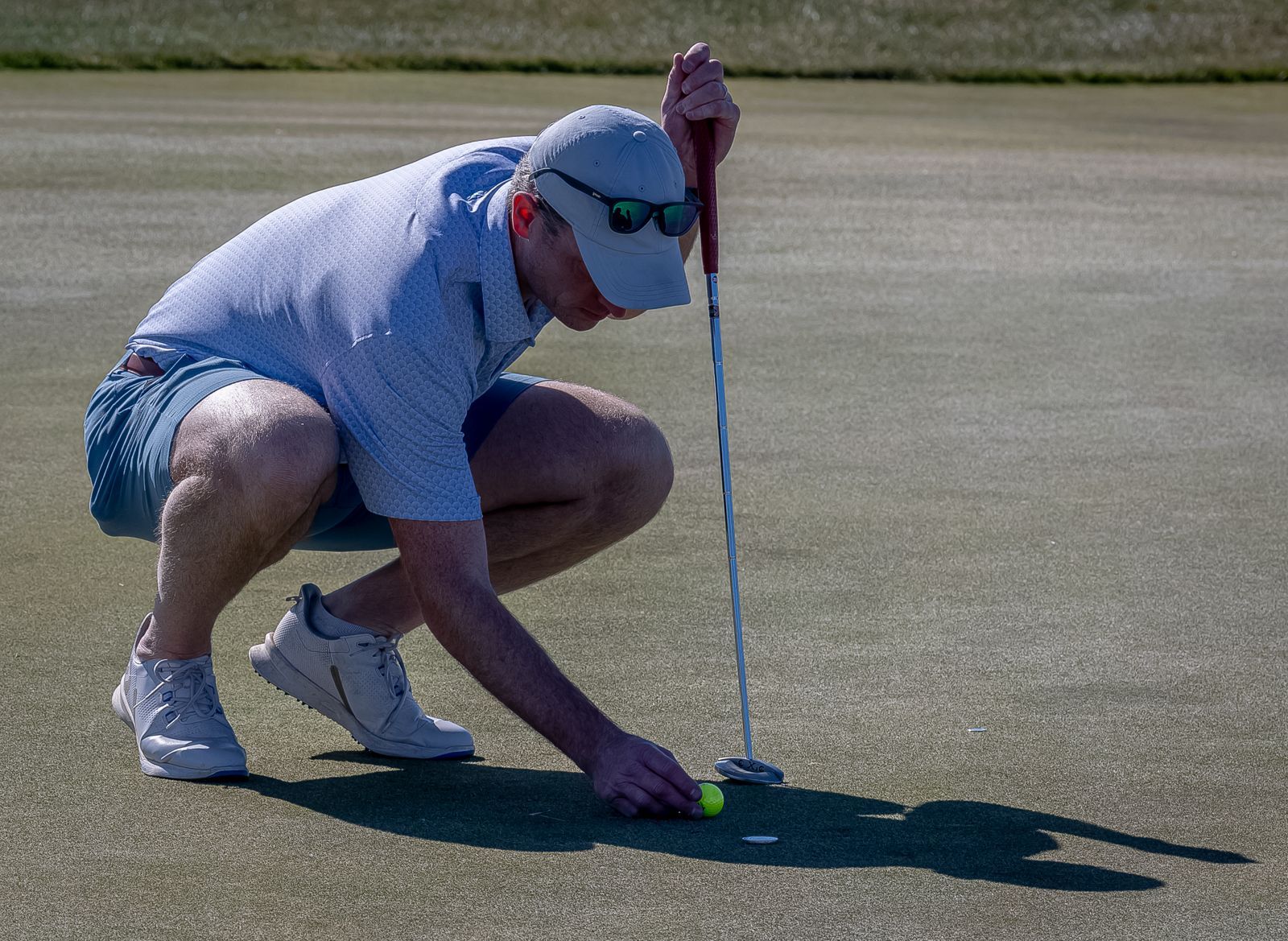 Golfer lines up a putt on a green. He crouches low, wearing sunglasses, cap, and light-colored clothing.