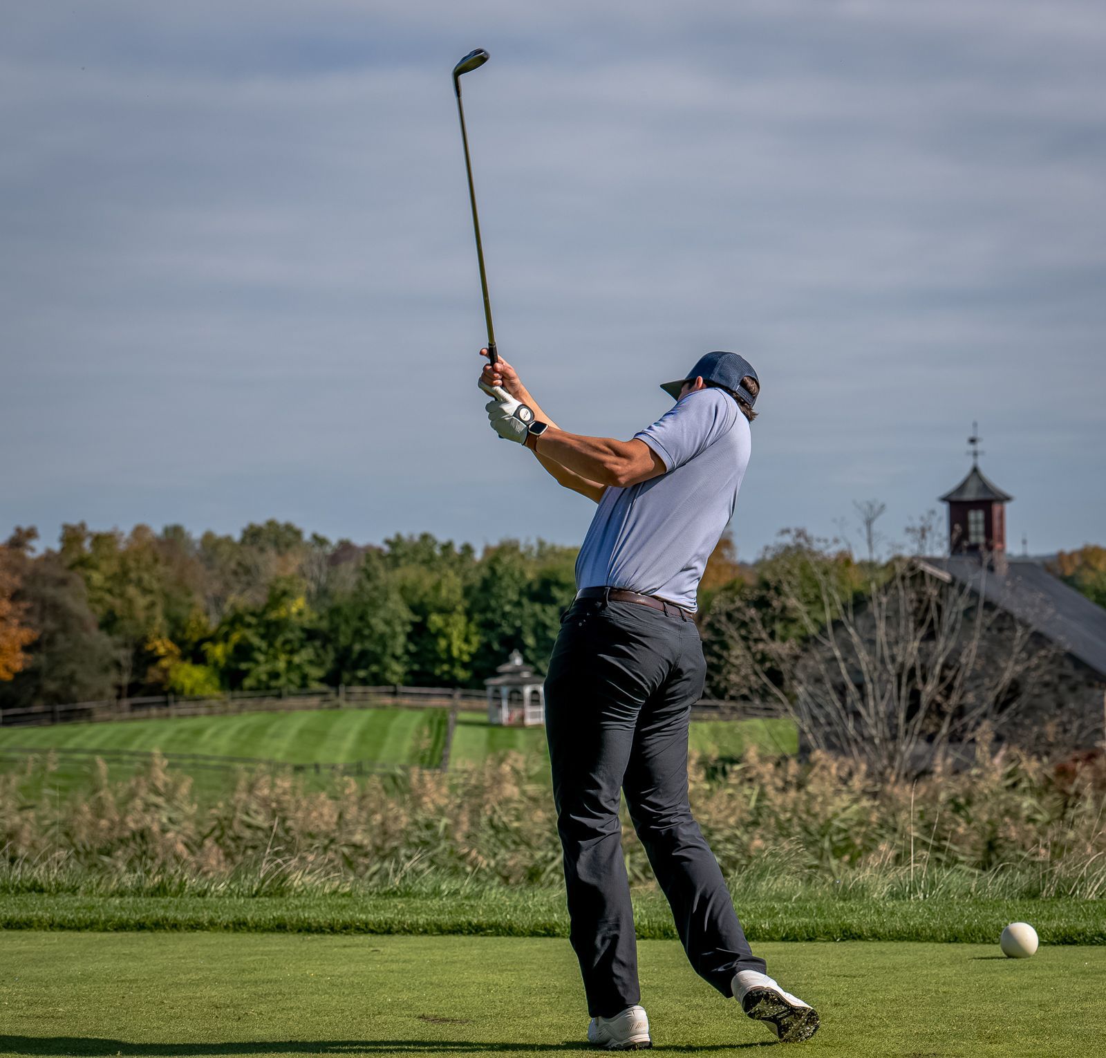Golfer swinging club on a golf course under a blue sky, near a barn and trees.