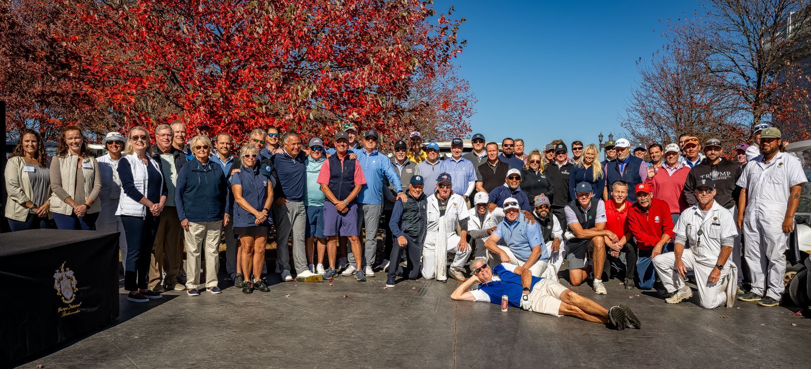 Group of people posing outdoors on a sunny day. Red fall foliage in the background. Some are kneeling or laying.