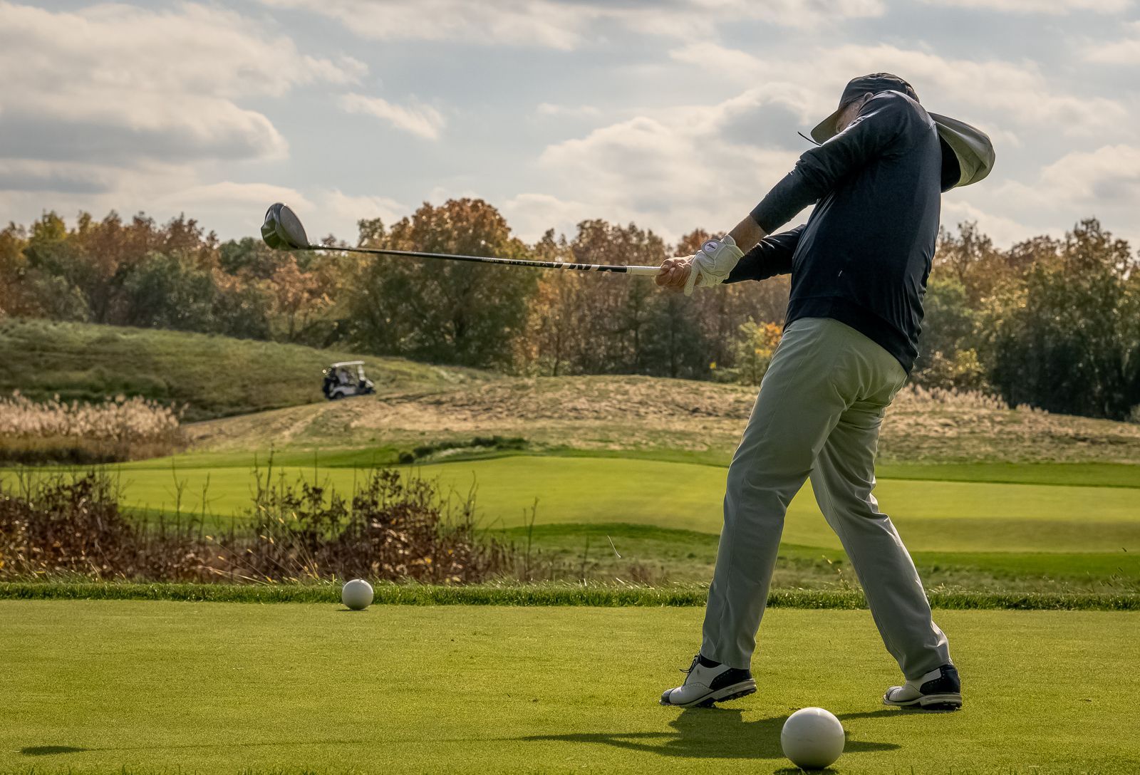 Golfer swinging a club on a green course, with a golf cart and trees in the background. Sunny day.