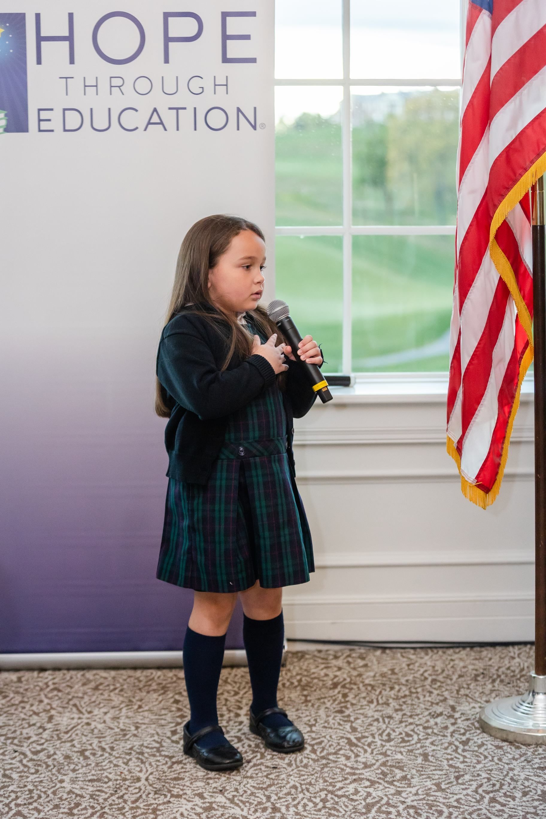 Young girl in plaid dress speaks at microphone next to American flag and banner that says 