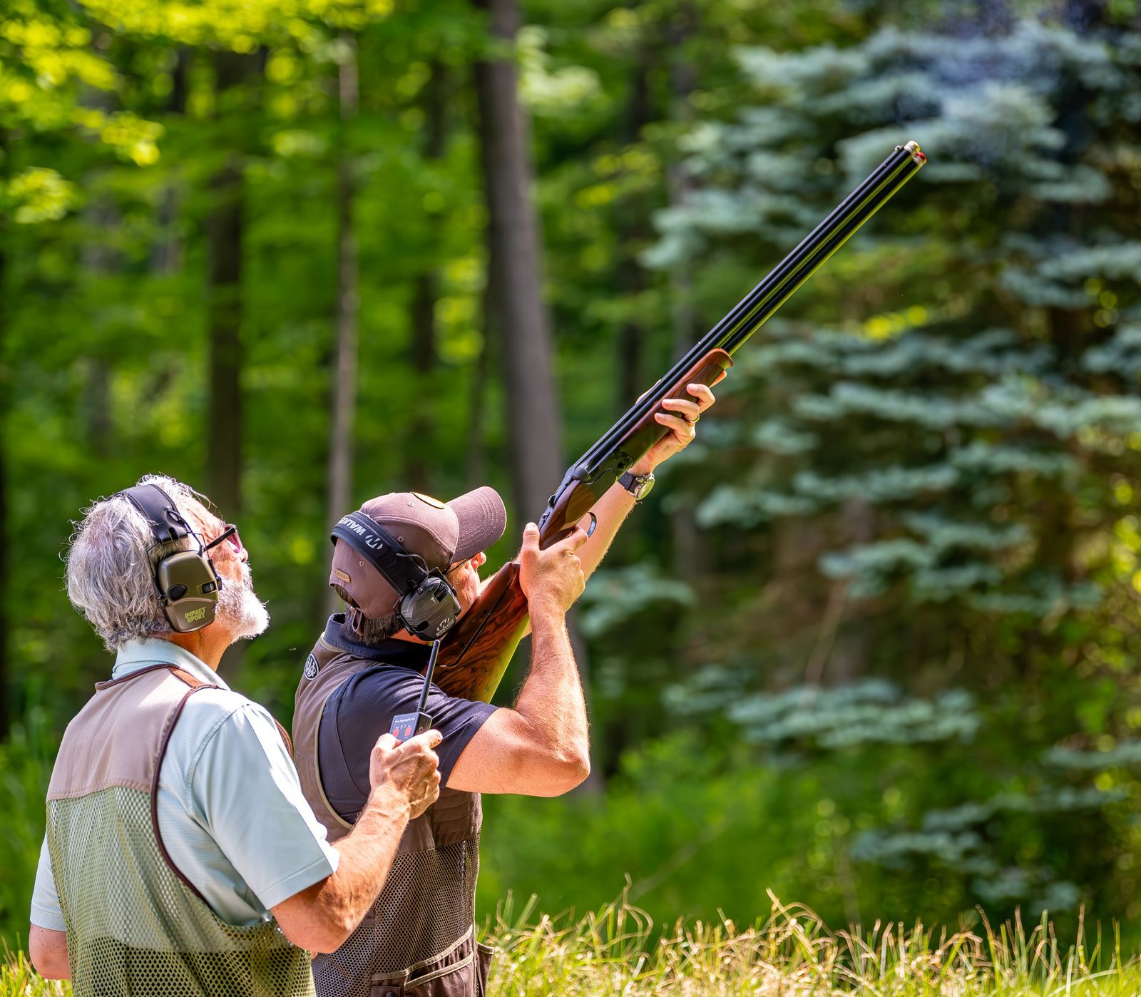 Two people shooting a shotgun outdoors, one aiming, the other observing. Green trees in background.