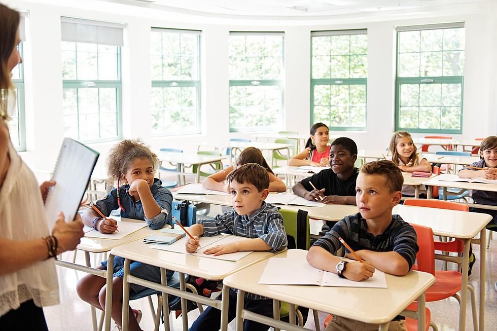 A teacher with a tablet in a bright classroom, instructing students at their desks.