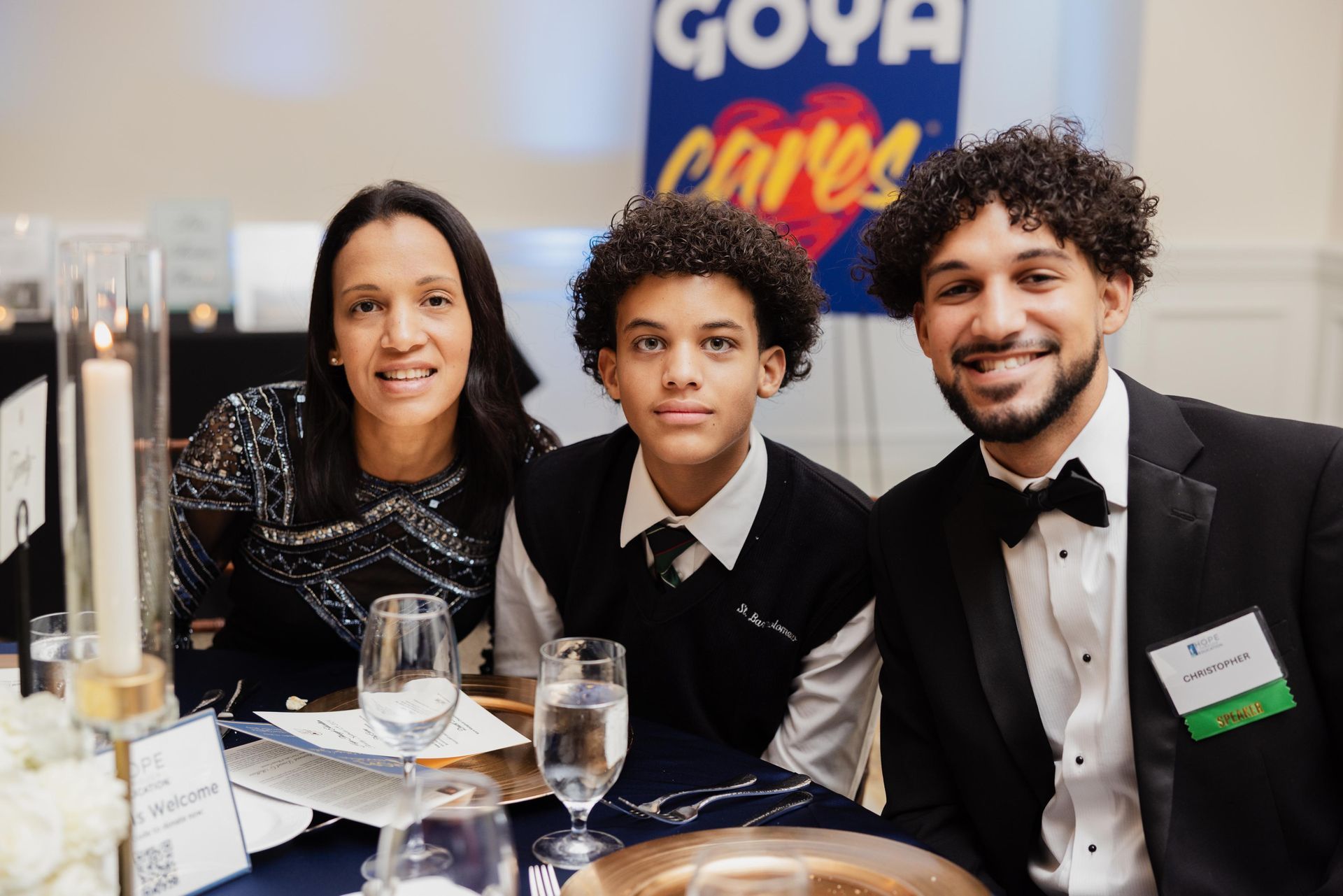 Family at an event with Goya Cares banner, smiling at the camera. Woman in sparkly top, boy in school uniform, man in tuxedo.