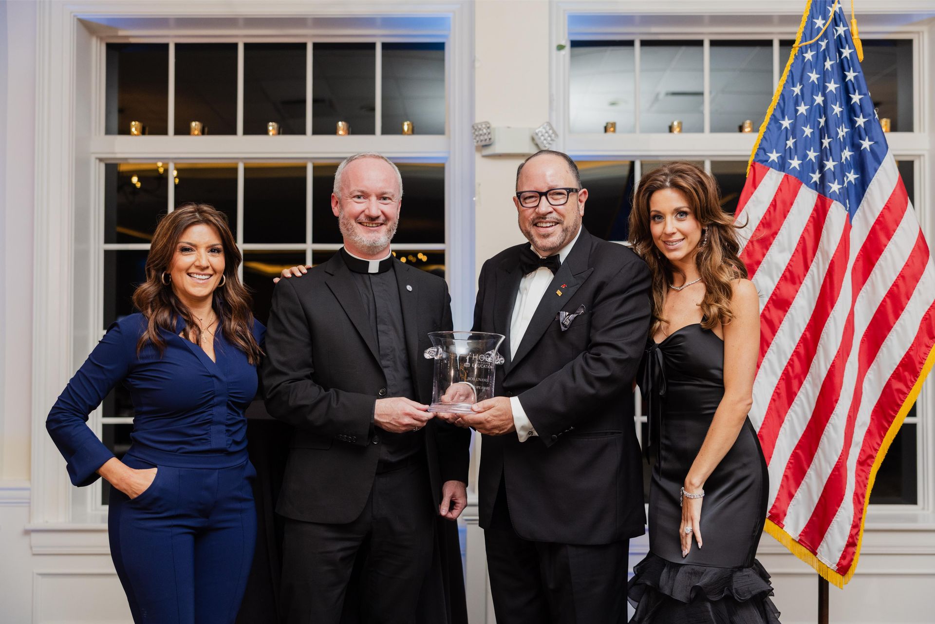 Four people holding award near American flag. Formal attire, smiling. Interior setting.
