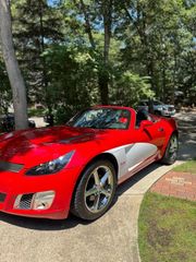 A bright red convertible sports car with a silver side panel parked on a driveway near trees on a sunny day.