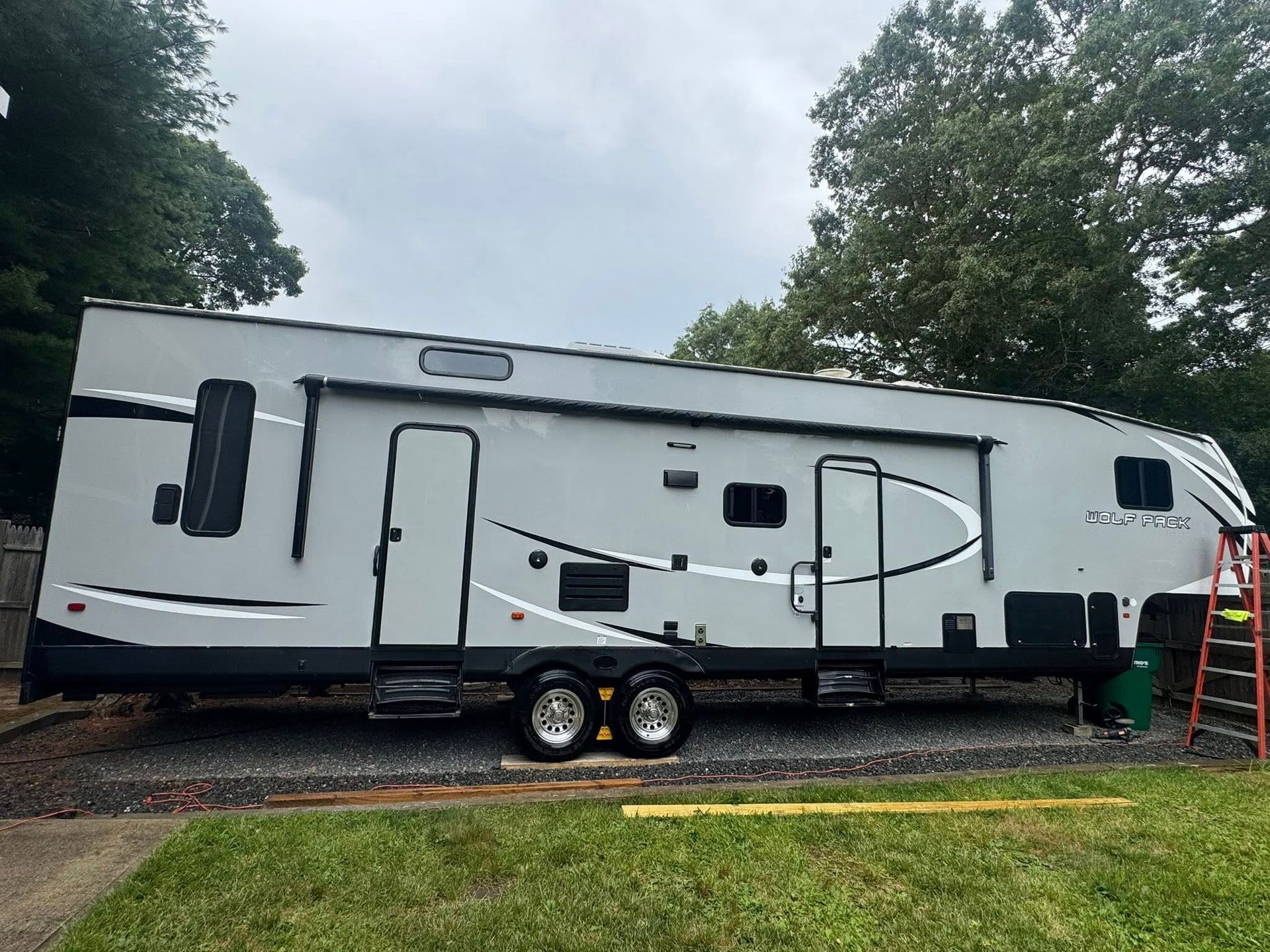 A light gray recreational vehicle parked on a gravel lot near a green lawn and trees, with a red ladder leaned against it.