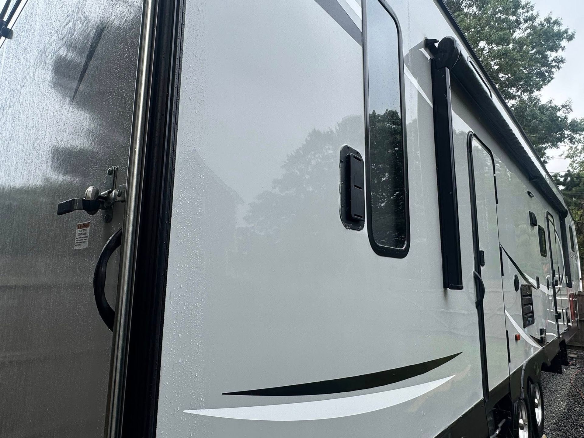 An angled, close-up exterior view of a light gray travel trailer with black window trim, decals, and an extended awning.