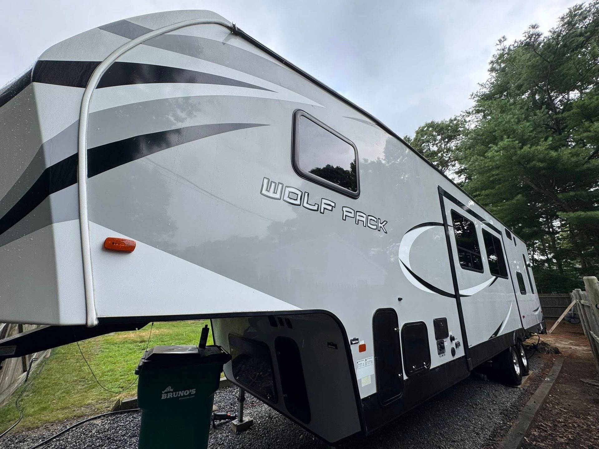 A white and gray Wolf Pack fifth-wheel camper parked on a gravel surface outdoors with trees in the background.