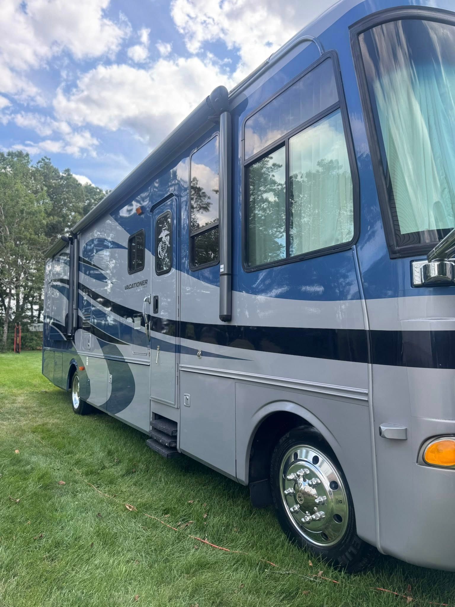 A large, modern RV with blue and silver paint parked on a grassy field under a cloudy, blue sky.