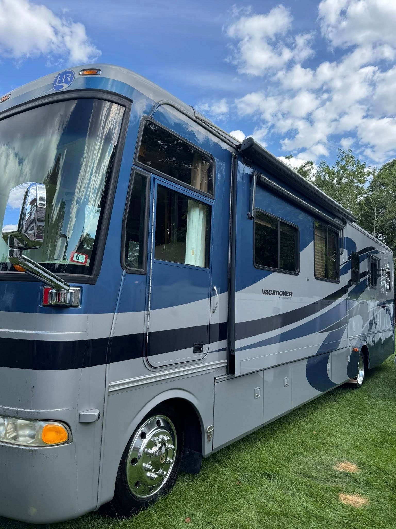 A blue and silver recreational vehicle parked on a grassy field under a sunny, cloudy sky.