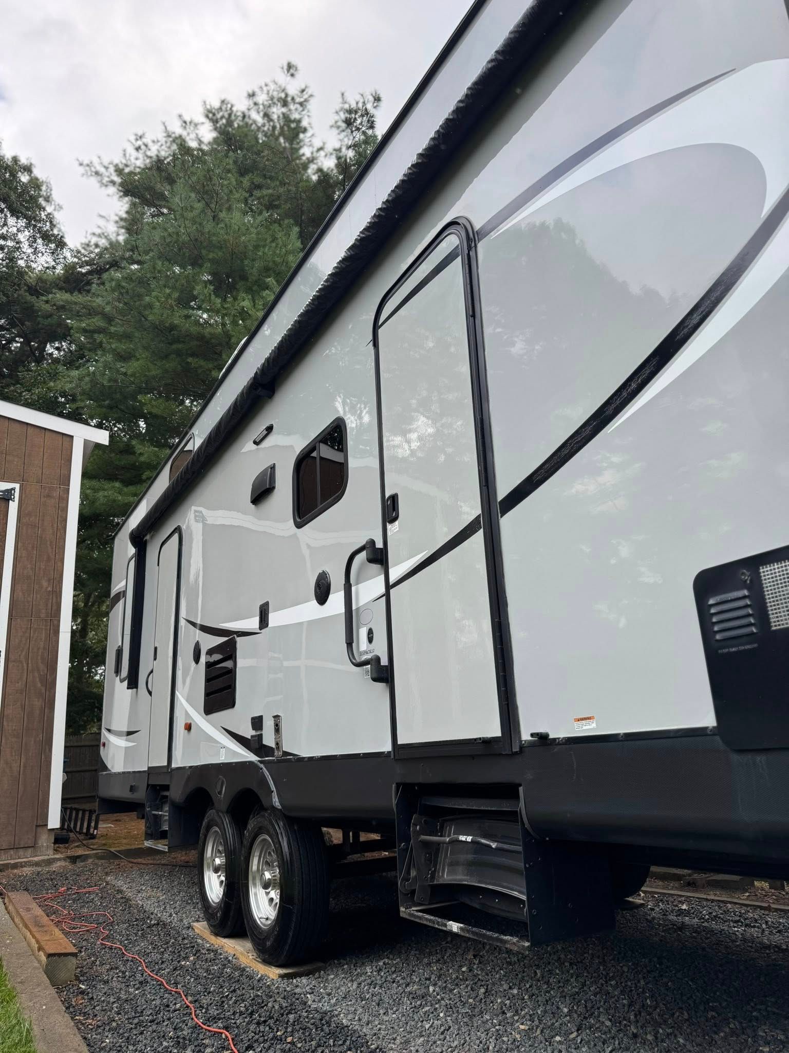 A side view of a light gray, tandem-axle travel trailer parked on gravel beside a small wooden structure.