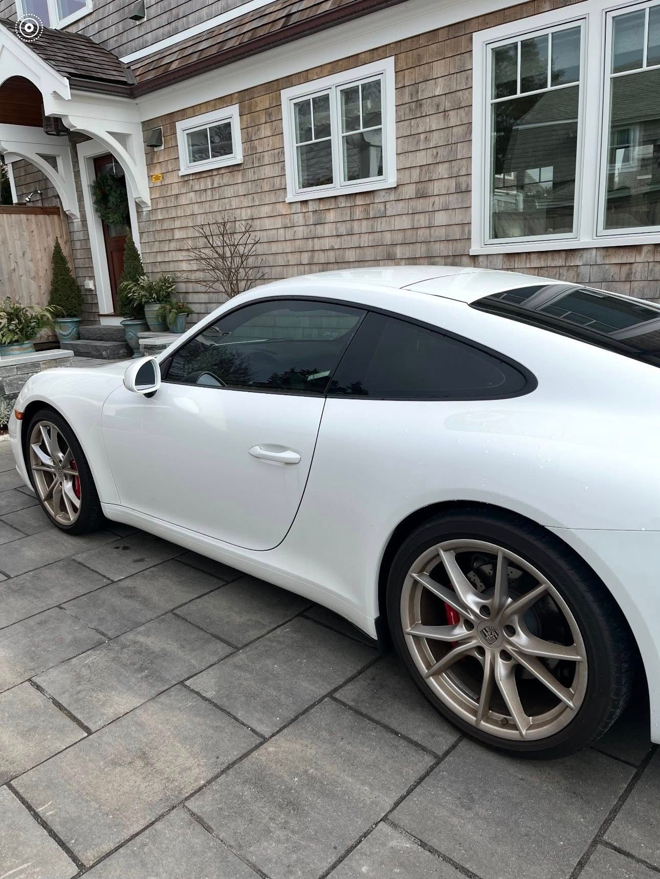 A white Porsche sports car parked on a paved driveway in front of a shingled house.