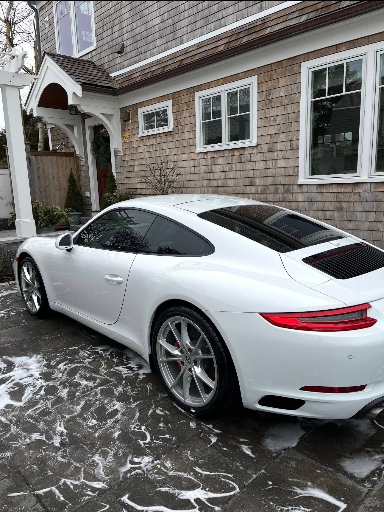 A white Porsche parked on a patio in front of a house with wood shingle siding and white trim.