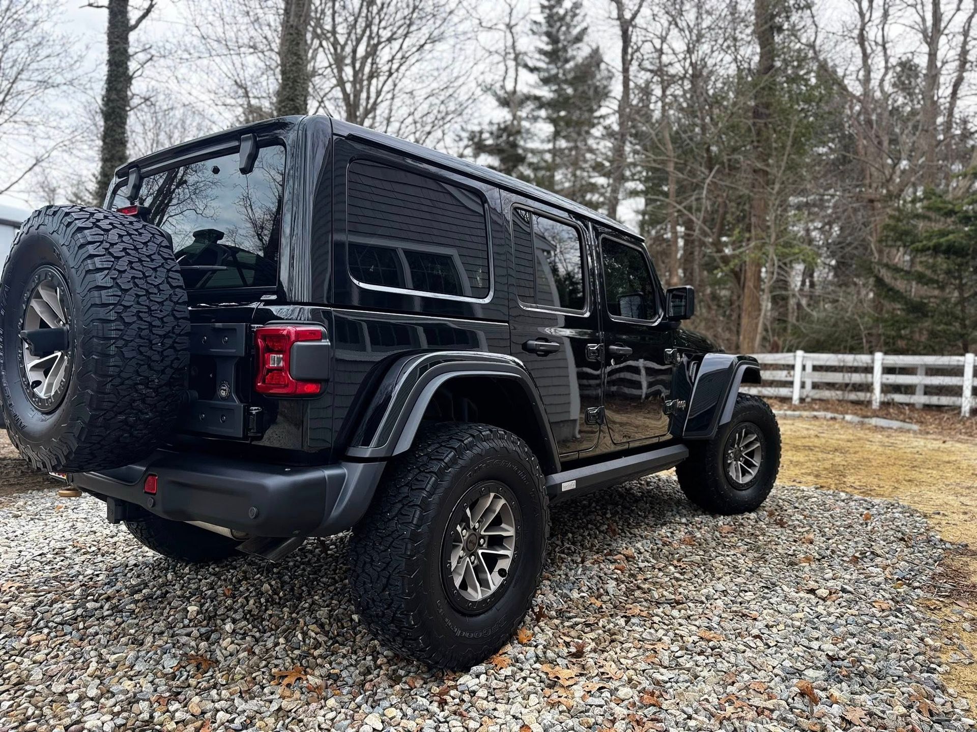 A black Jeep Wrangler parked on a gravel driveway with a sparse wooded area in the background.
