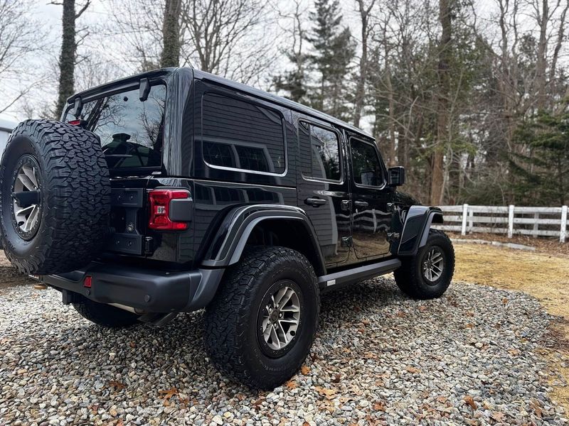 A black Jeep Wrangler parked on a gravel driveway with a sparse wooded area in the background.
