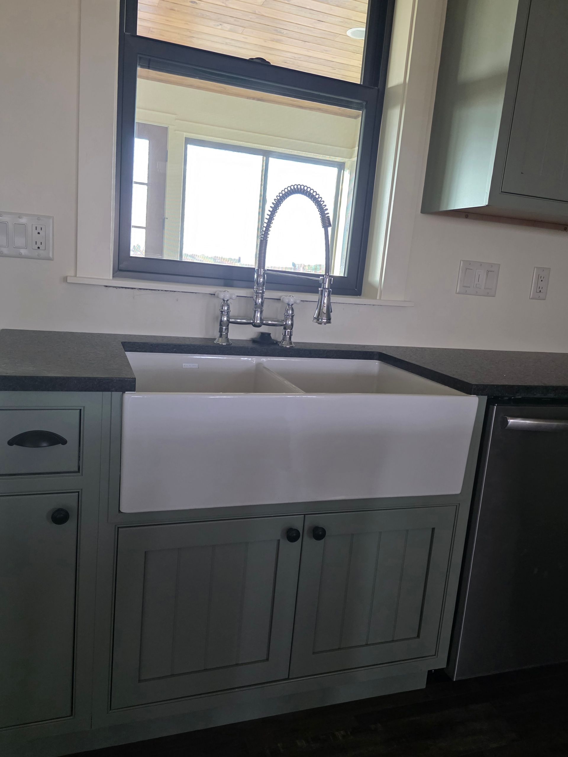Kitchen sink with white apron-front basin, gray cabinets, and a window above the faucet.