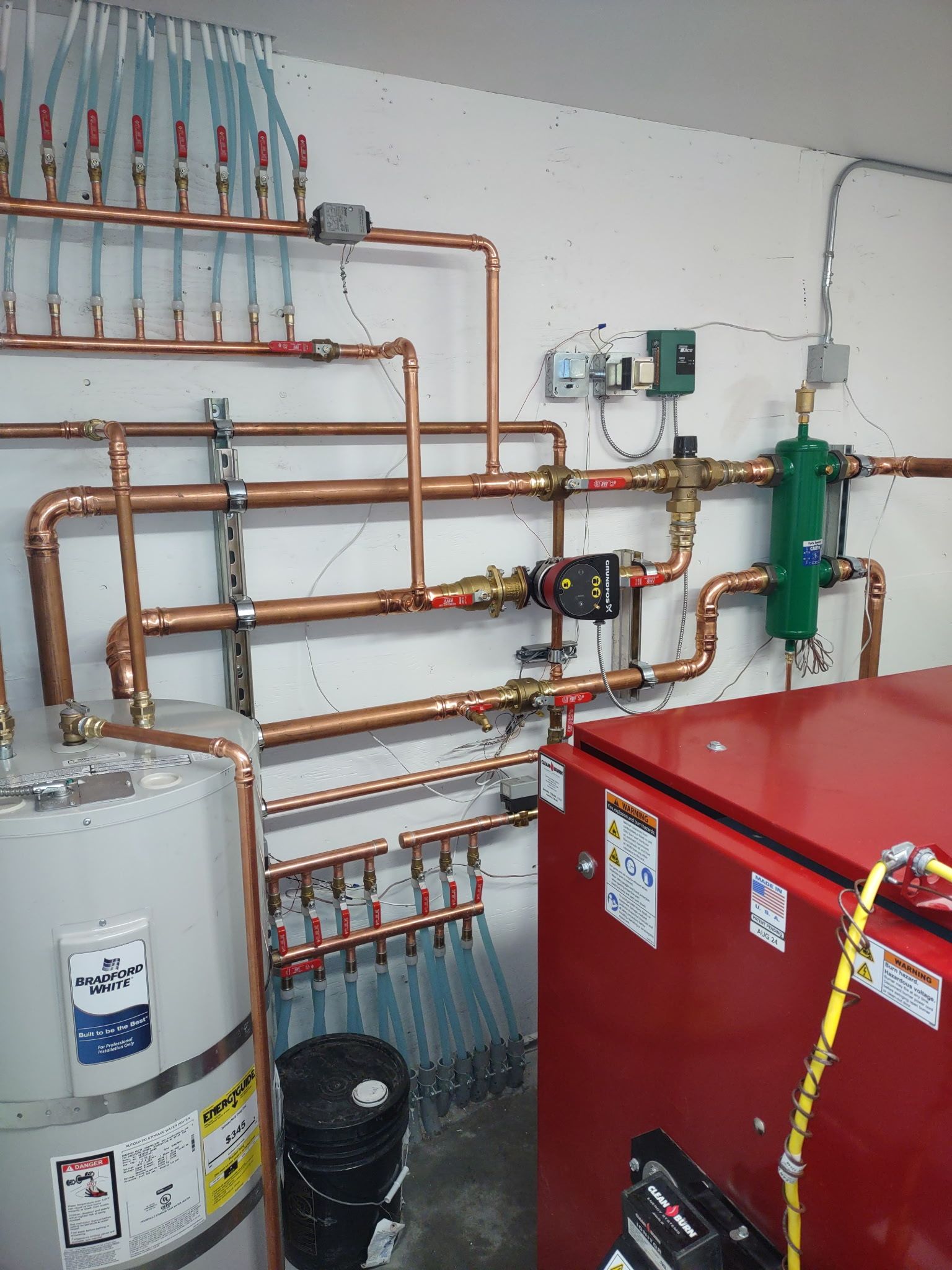 Boiler room with copper piping, white tank, and red equipment against a white wall