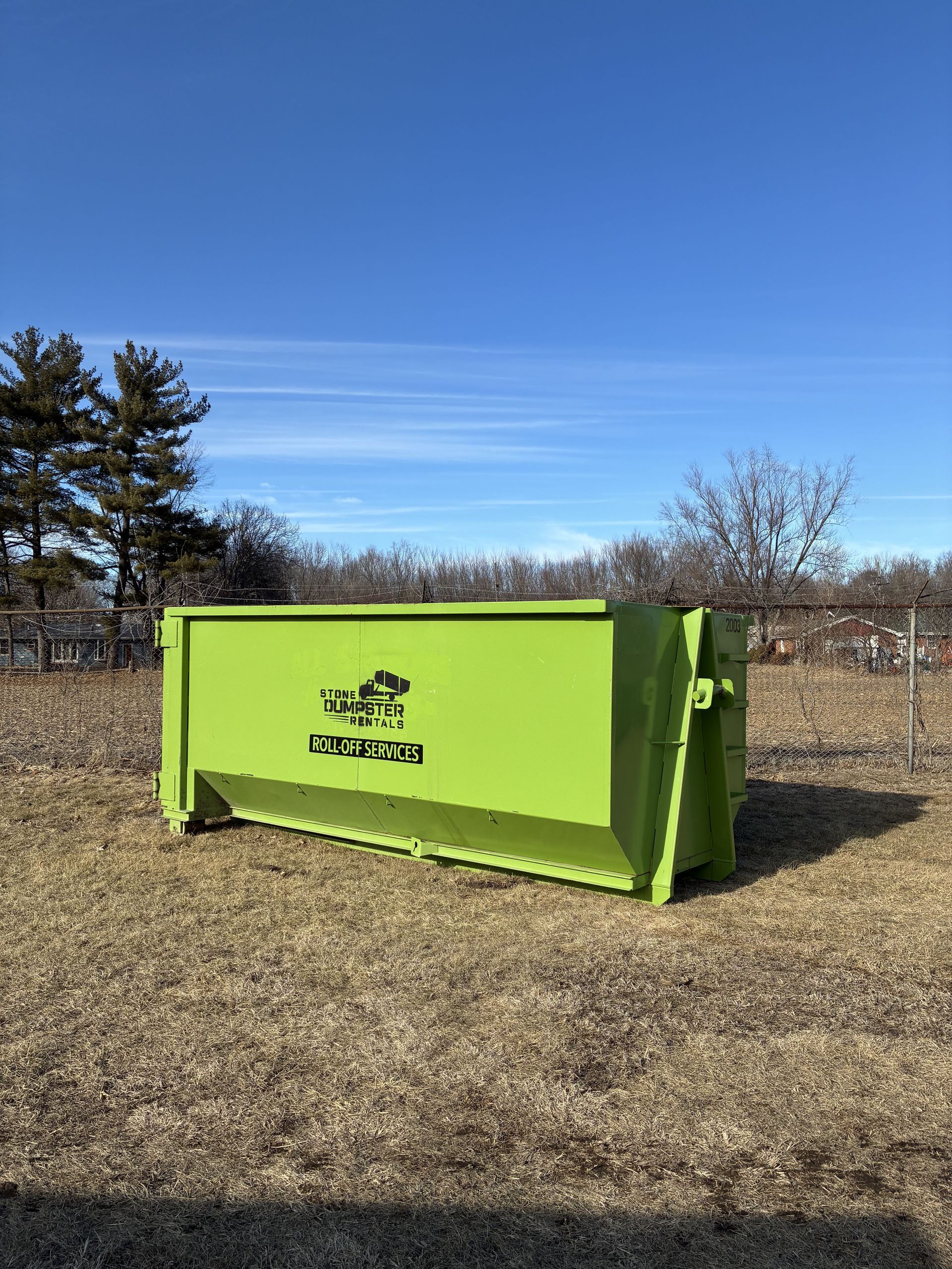 A large, brown, rusted roll-off dumpster filled with various construction debris, parked on a concrete path near grass. A large, brown, rusted roll-off dumpster filled with various construction debris, parked on a concrete path near grass.