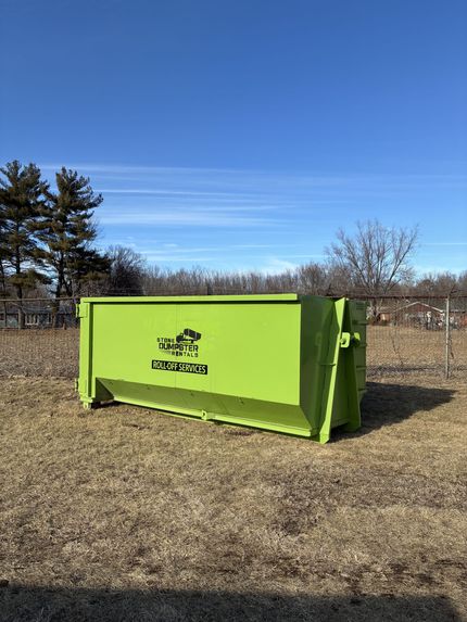 A large, brown, rusted roll-off dumpster filled with various construction debris, parked on a concrete path near grass.