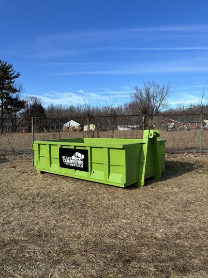 A blue metal dumpster sits on a paved driveway in front of a white house with trees in the yard.