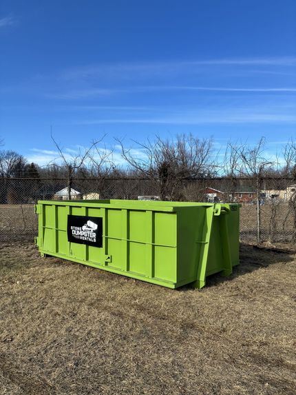 A large, empty blue dumpster sits on a dirt lot in front of a modern residential home under construction.