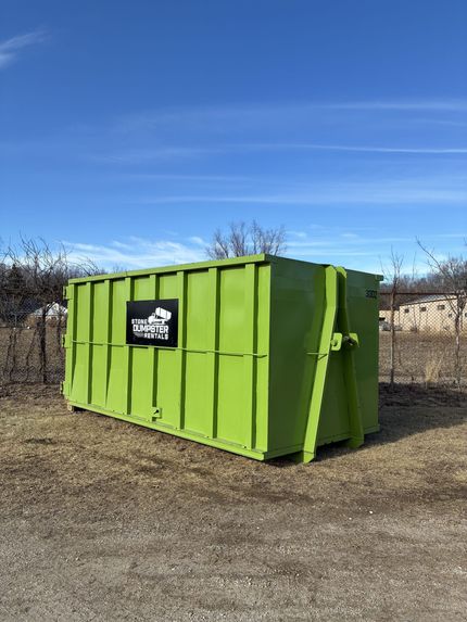 A large, empty green roll-off dumpster sitting on a paved surface with bare trees in the background.