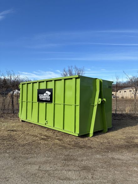 A large, empty green roll-off dumpster sitting on a paved surface with bare trees in the background.