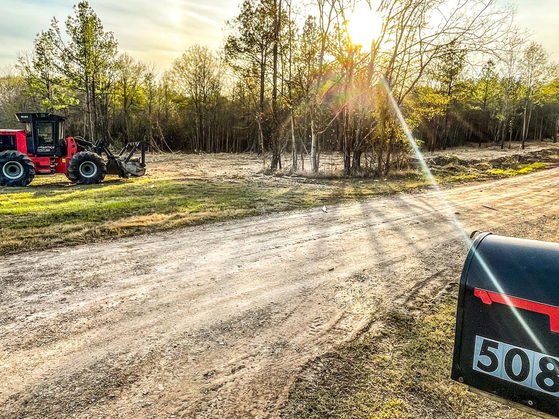 Sunlit dirt road beside a wooded field, with a red tractor on the left and a mailbox in the foreground.