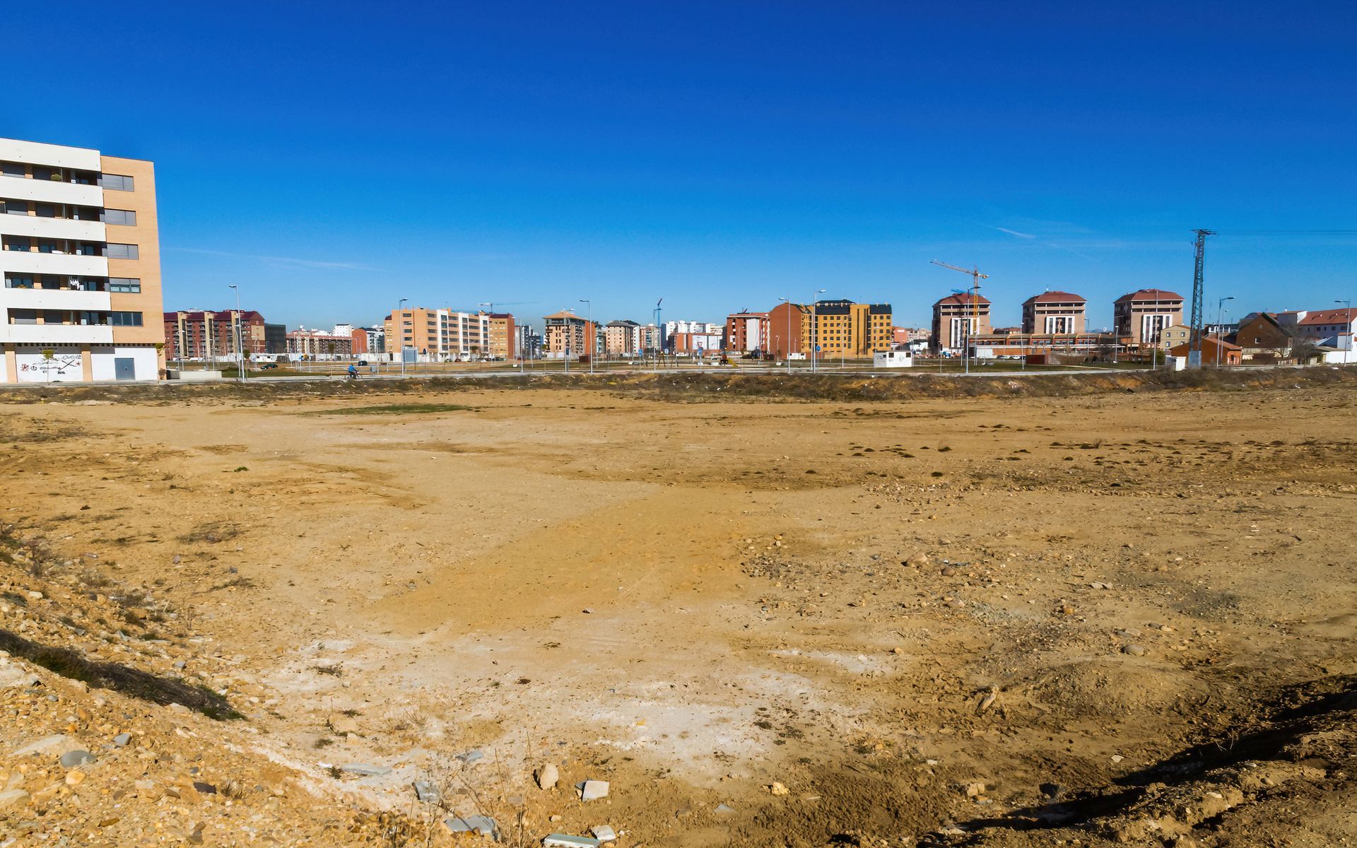 Empty dirt lot under a clear blue sky, with apartment buildings in the distance.